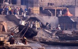 A group of mourners surround a body prior to cremation. In the foreground two Doms sift through the ashes of a previous cremation