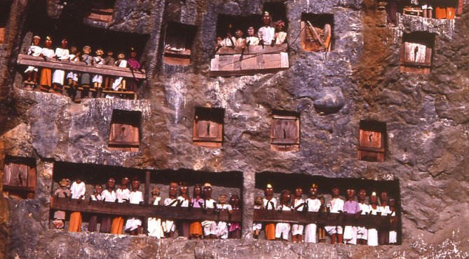 Tau tau watch over the village of Lemo from one of many family balconies. The square wooden covers are family vaults housing the skeletal remains of the dead. The freshness of the clothing and the deep tones of the wood on all the effigies suggest they have recently been replaced. Having great value to museums and private collectors, many Tau tau are either stolen or sold off over time. 